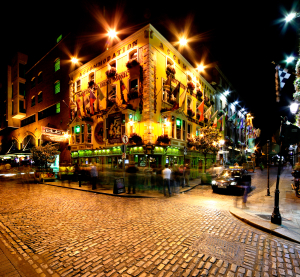 Ein traditioneller irischer Pub in Dublin bei Nacht, hell erleuchtet mit bunten Fassaden und Fahnen, Menschen flanieren &uuml;ber das Kopfsteinpflaster.