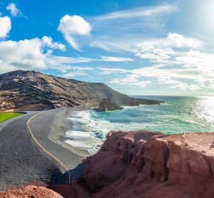 K&uuml;stenstra&szlig;e auf Lanzarote mit schwarzem Lavastrand, r&ouml;tlichen Felsen und Blick auf den Atlantik unter blauem Himmel.