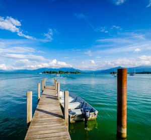Holzsteg mit Boot auf einem ruhigen See in Bayern, im Hintergrund eine kleine Insel und die Alpen unter blauem Himmel mit wei&szlig;en Wolken.