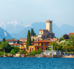 Blick auf das mittelalterliche Castello Scaligero in Malcesine am Gardasee, umgeben von bunten H&auml;usern, mit den Alpen im Hintergrund und blauem Wasser im Vordergrund.