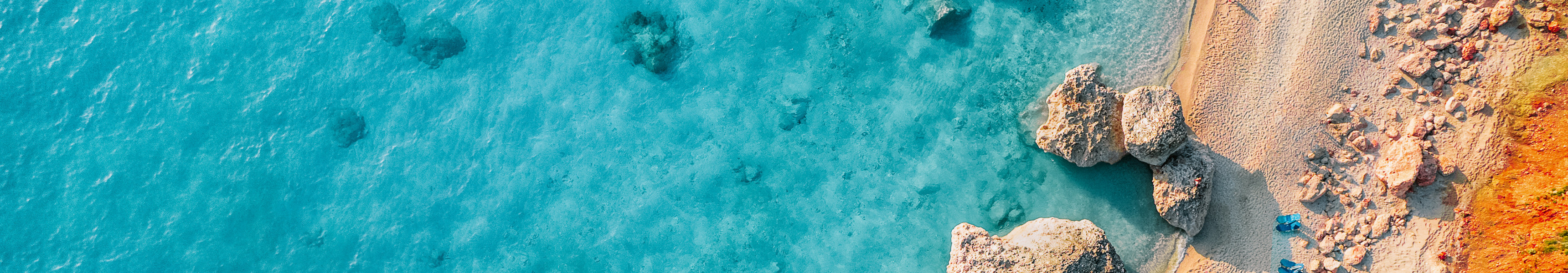 Drohnenaufnahme eines Strandes mit türkisblauem Wasser, Felsen im Sand und Reihen von Liegestühlen.