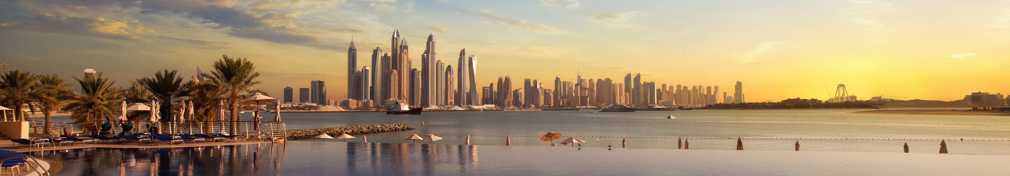 Infinity-Pool mit Palmen und Sonnenliegen im Vordergrund, dahinter das Meer und die Skyline von Dubai mit ihren modernen Hochh&auml;usern bei Sonnenuntergang.
