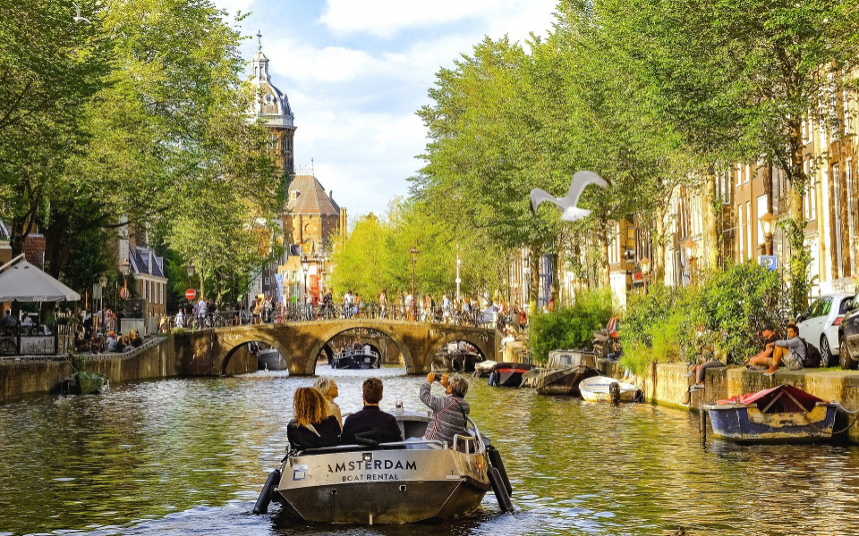 Boot mit Touristen auf einer Grachtenrundfahrt durch das gr&uuml;ne Kanalnetz von Amsterdam mit Blick auf eine historische Br&uuml;cke und Kirchturm.