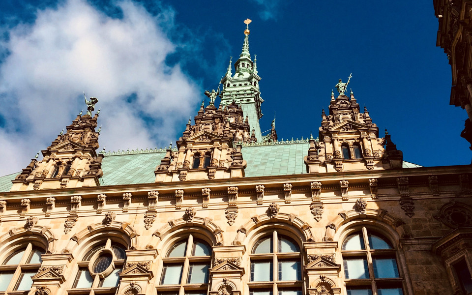Hamburger Rathaus, ein pr&auml;chtiges Geb&auml;ude im Stil der Neorenaissance mit verzierten Fassaden, gro&szlig;en Fenstern und einem hohen gr&uuml;nen Turm mit vergoldeter Spitze vor blauem Himmel.