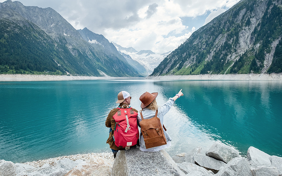 Zwei Wanderer mit Rucks&auml;cken sitzen am Ufer eines t&uuml;rkisfarbenen Bergsees inmitten der Alpen.