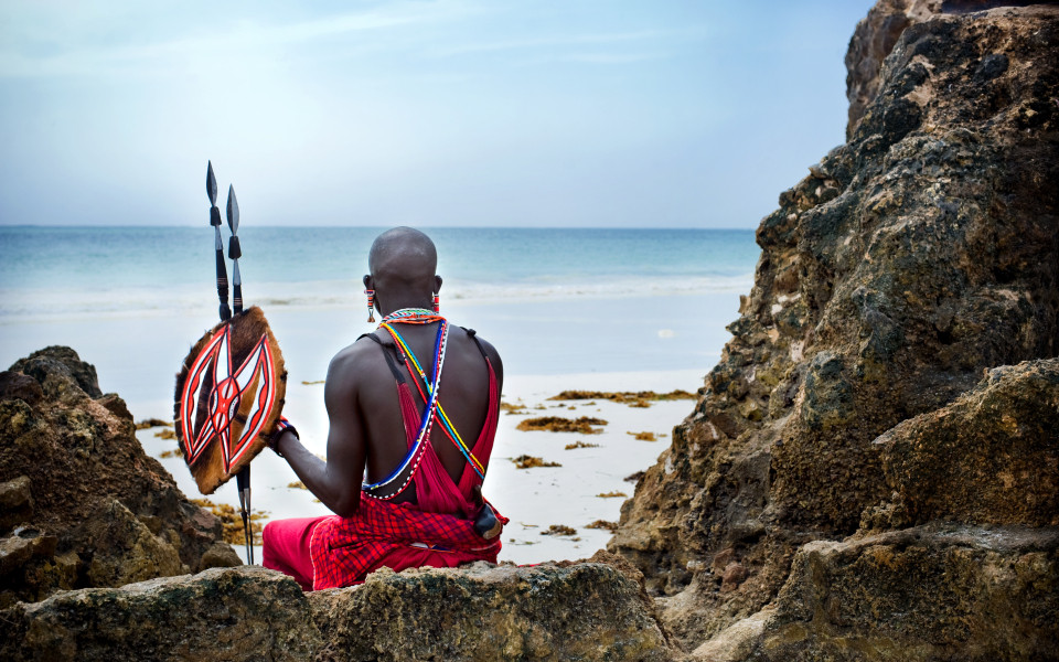 Ein Maasai sitzt auf einem Felsen am Strand von Kenia und blickt auf das Meer.