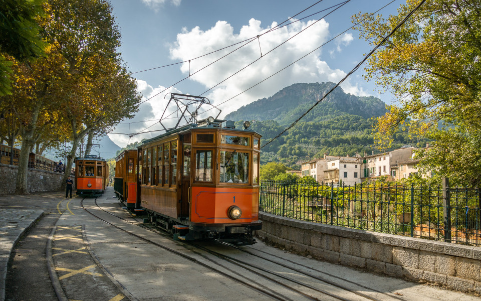 Historische Stra&szlig;enbahn &bdquo;Roter Blitz&ldquo; auf Mallorca, umgeben von gr&uuml;nen Bergen, malerischen D&ouml;rfern und mediterraner Landschaft.