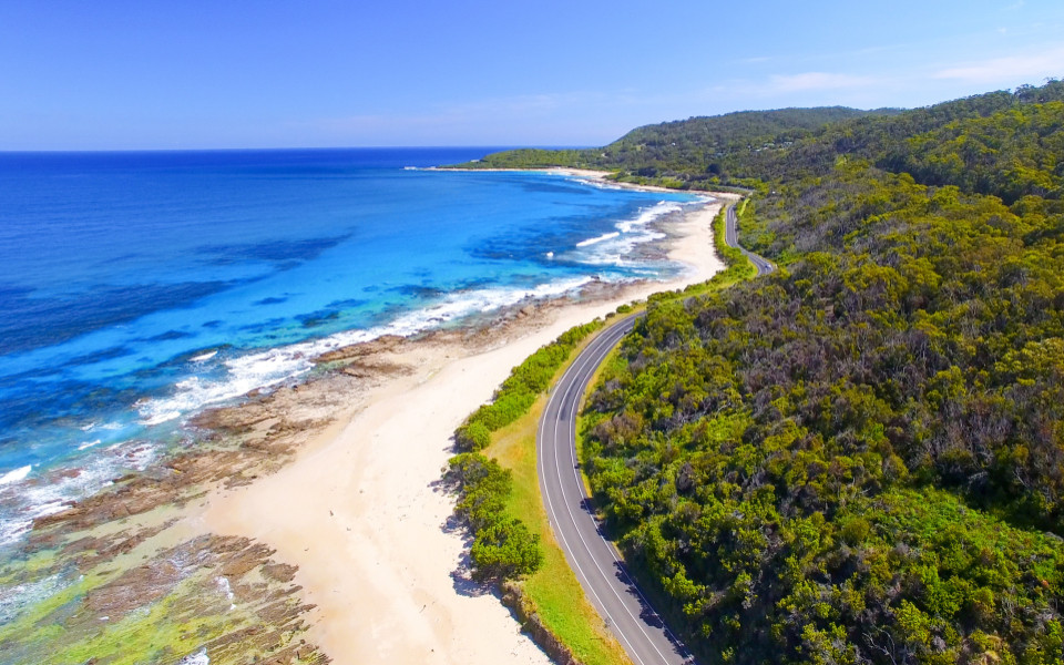 Stra&szlig;enverlauf der Great Ocean Road zwischen bewaldetem Hang und felsiger K&uuml;ste mit Blick auf den Ozean.
