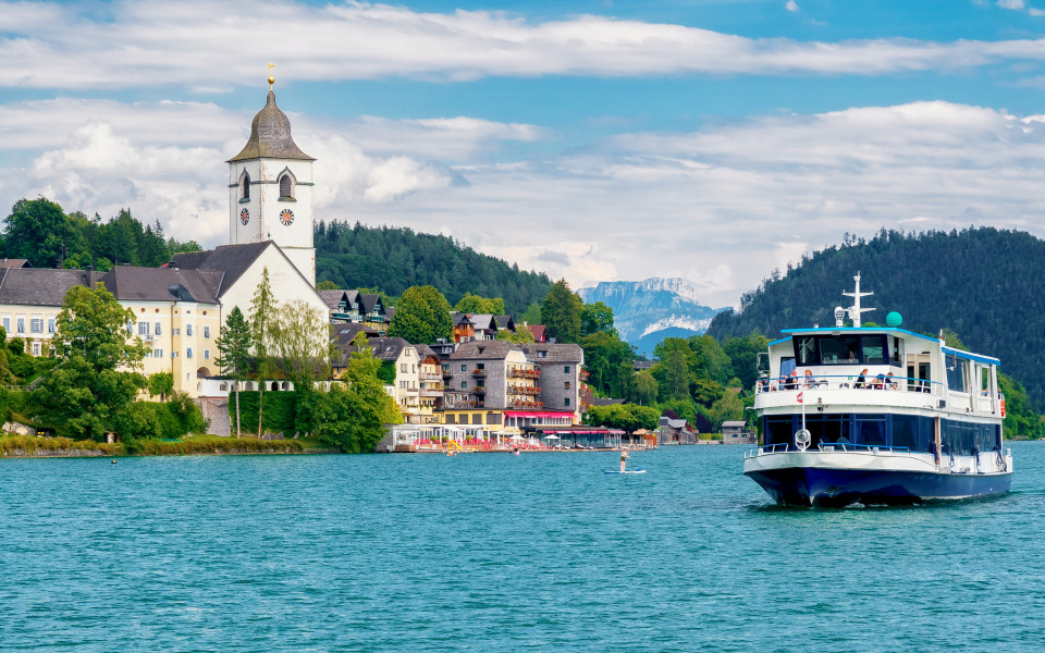 Der Wolfgangsee mit einem Ausflugsboot im Vordergrund und einer malerischen Landschaft, einschließlich der markanten Kirche und der Berge im Hintergrund.