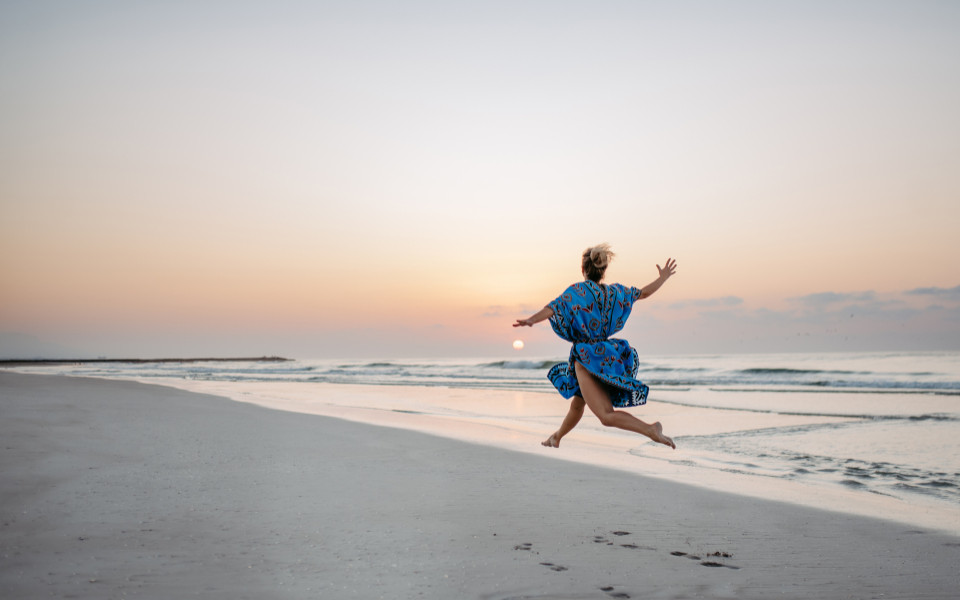 Junge Frau genießt die Zeit am Strand und springt. Im Hintergrund sieht man das Meer mit Sonnenuntergang.