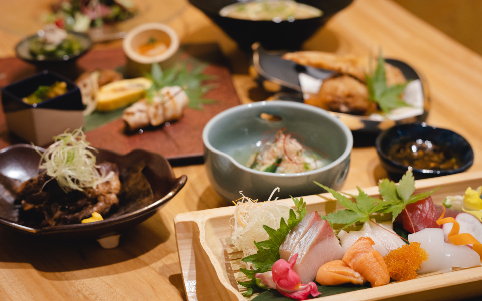 Japanisches Men&uuml; auf einem Holztisch mit kunstvoll angerichtetem Sashimi (Lachs, Thunfisch, wei&szlig;er Fisch und Ikura) auf einem Holzbrett, dazu mehrere kleine Schalen mit Beilagen, Suppe und frittierten Speisen im Hintergrund.